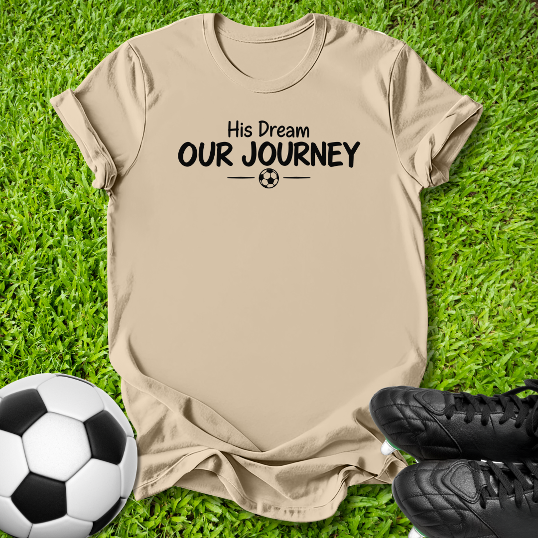 beige short-sleeve t-shirt with the black printed text 'His Dream OUR JOURNEY' and a small soccer ball graphic, laid flat on green grass with a soccer ball and black cleats nearby.