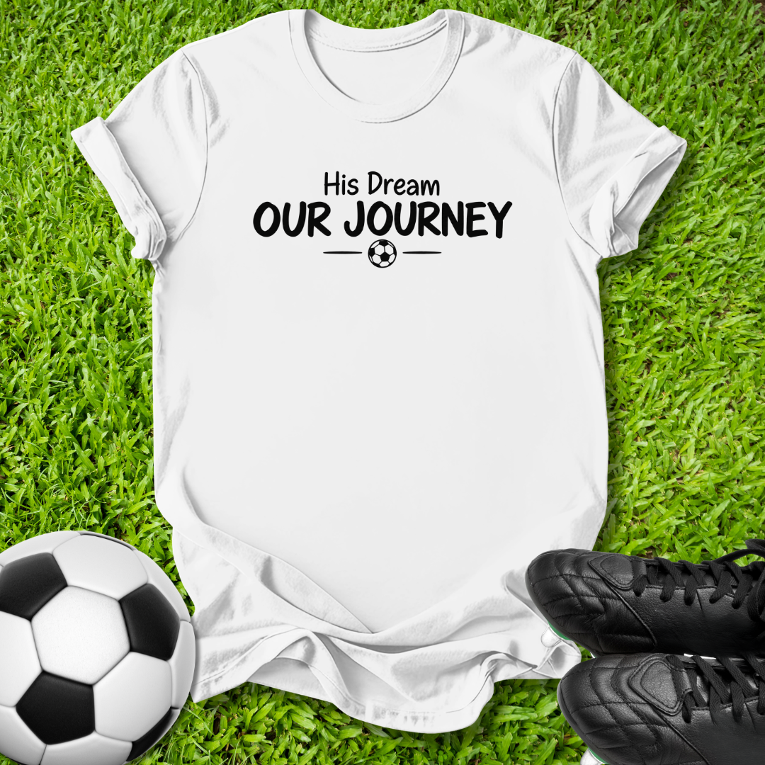 A white short-sleeve t-shirt with the black printed text 'His Dream OUR JOURNEY' and a small soccer ball graphic, laid flat on green grass with a soccer ball and black cleats nearby.