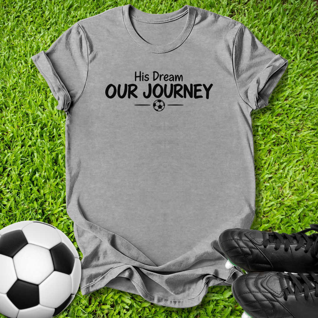 A heather gray short-sleeve t-shirt with the black printed text 'Her Dream OUR JOURNEY' and a small soccer ball graphic, laid flat on green grass with a soccer ball and black cleats as props.