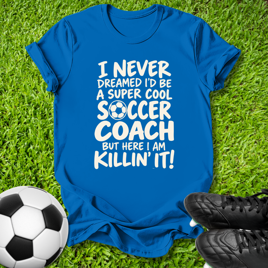 A royal blue short-sleeve t-shirt with a white graphic text that reads 'I NEVER DREAMED I'D BE A SUPER COOL SOCCER COACH BUT HERE I AM KILLIN' IT!' featuring a soccer ball icon inside the letter O. The shirt is laid flat on green grass with a soccer ball on the bottom left and black cleats on the bottom right.