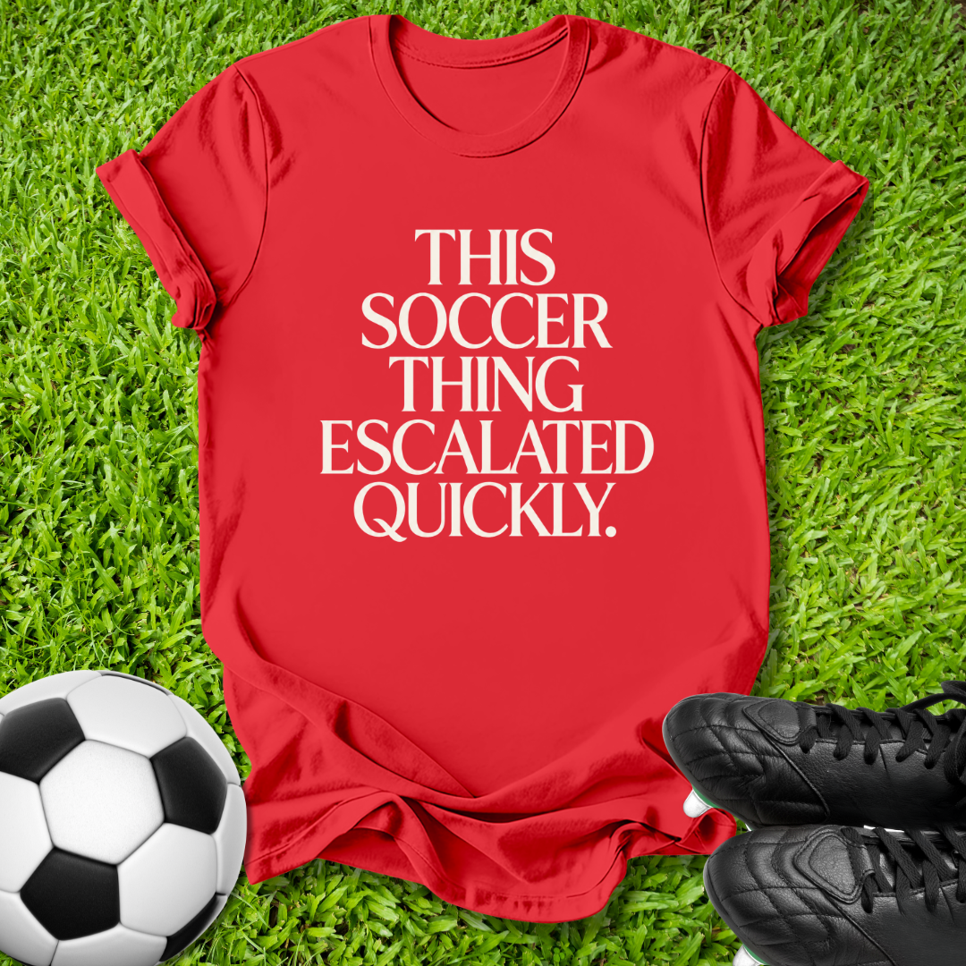 A red short-sleeve graphic T-shirt laid flat on green grass, with a soccer ball at the lower left and black cleats at the lower right. The shirt has large white text reading, 'THIS SOCCER THING ESCALATED QUICKLY.