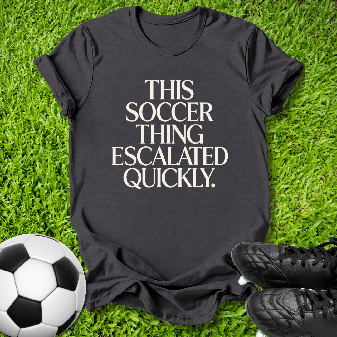 A black graphic t-shirt laid flat on green grass, featuring the text 'THIS SOCCER THING ESCALATED QUICKLY.' in white.