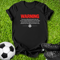 Black t-shirt with a humorous soccer warning graphic, laid flat on a green grass background with a soccer ball and black cleats as props.
