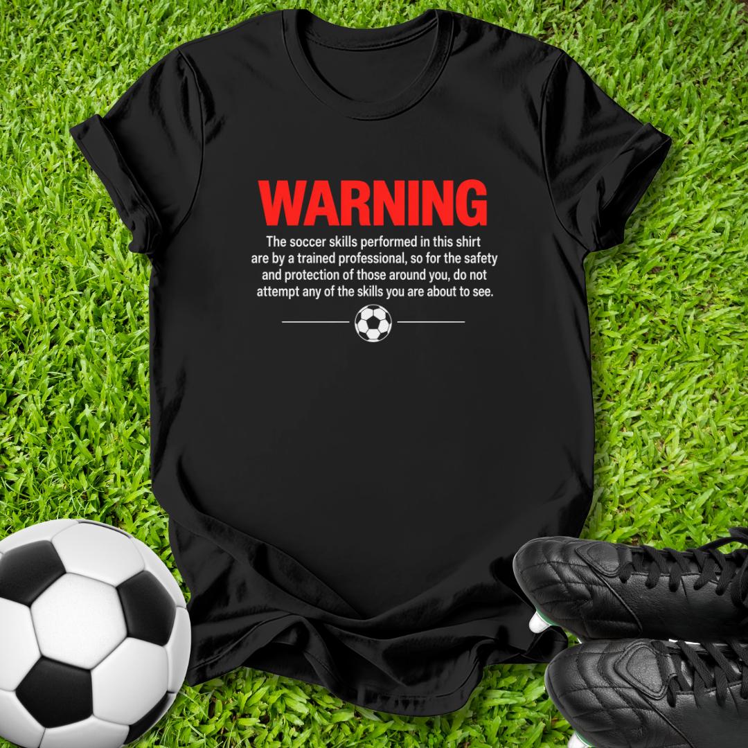 Black t-shirt with a humorous soccer warning graphic, laid flat on a green grass background with a soccer ball and black cleats as props.