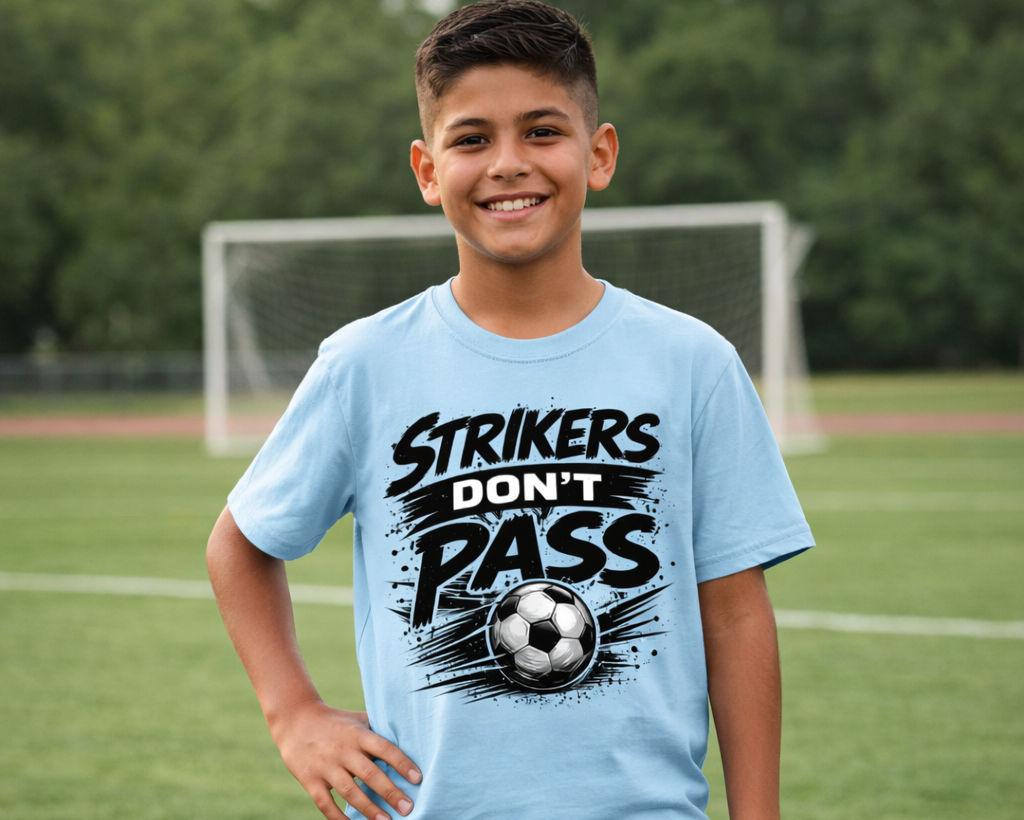 Young boy wearing a light blue t-shirt with a soccer-themed design on a grassy field.