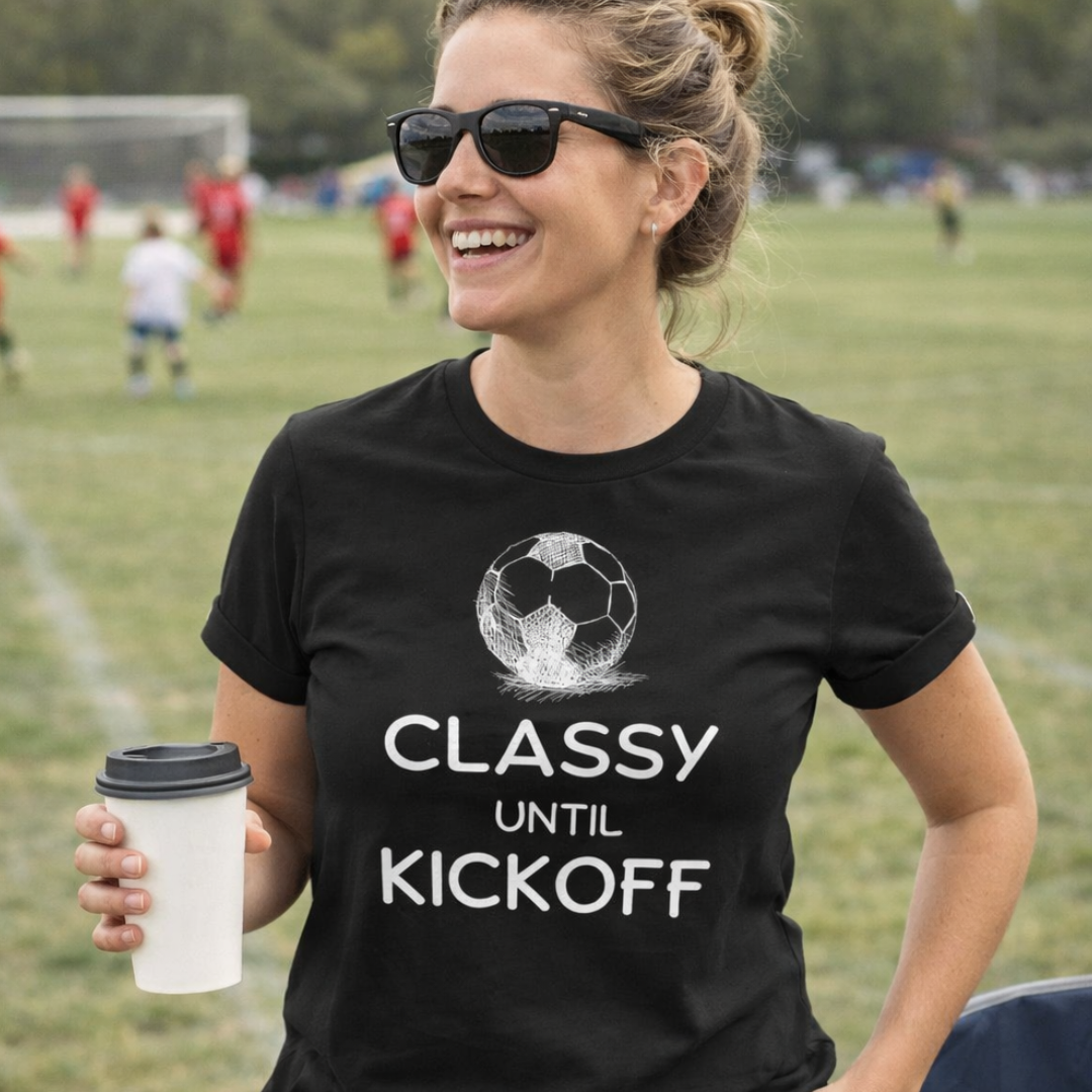 Woman wearing a black t-shirt with a soccer design and text, holding a coffee cup on a sports field.